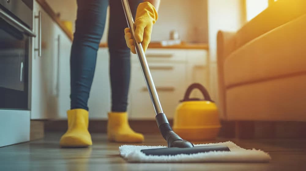 Person in yellow gloves and boots mopping a kitchen floor with a vacuum cleaner nearby.